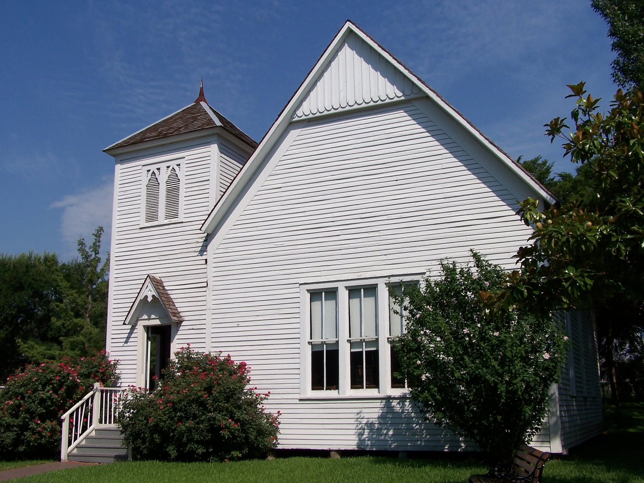 A large white wooden church with a steep roof and tower, surrounded by green grass and trees.